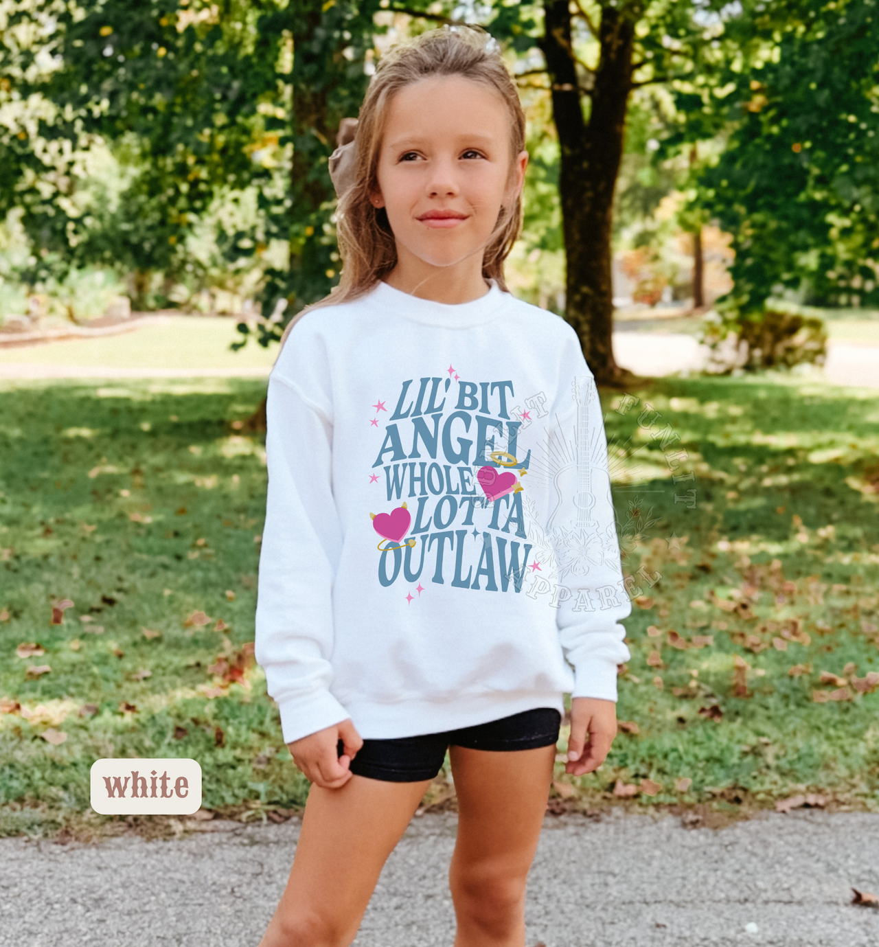 A young girl wearing a white sweatshirt with text printed on it, standing in a park surrounded by trees.