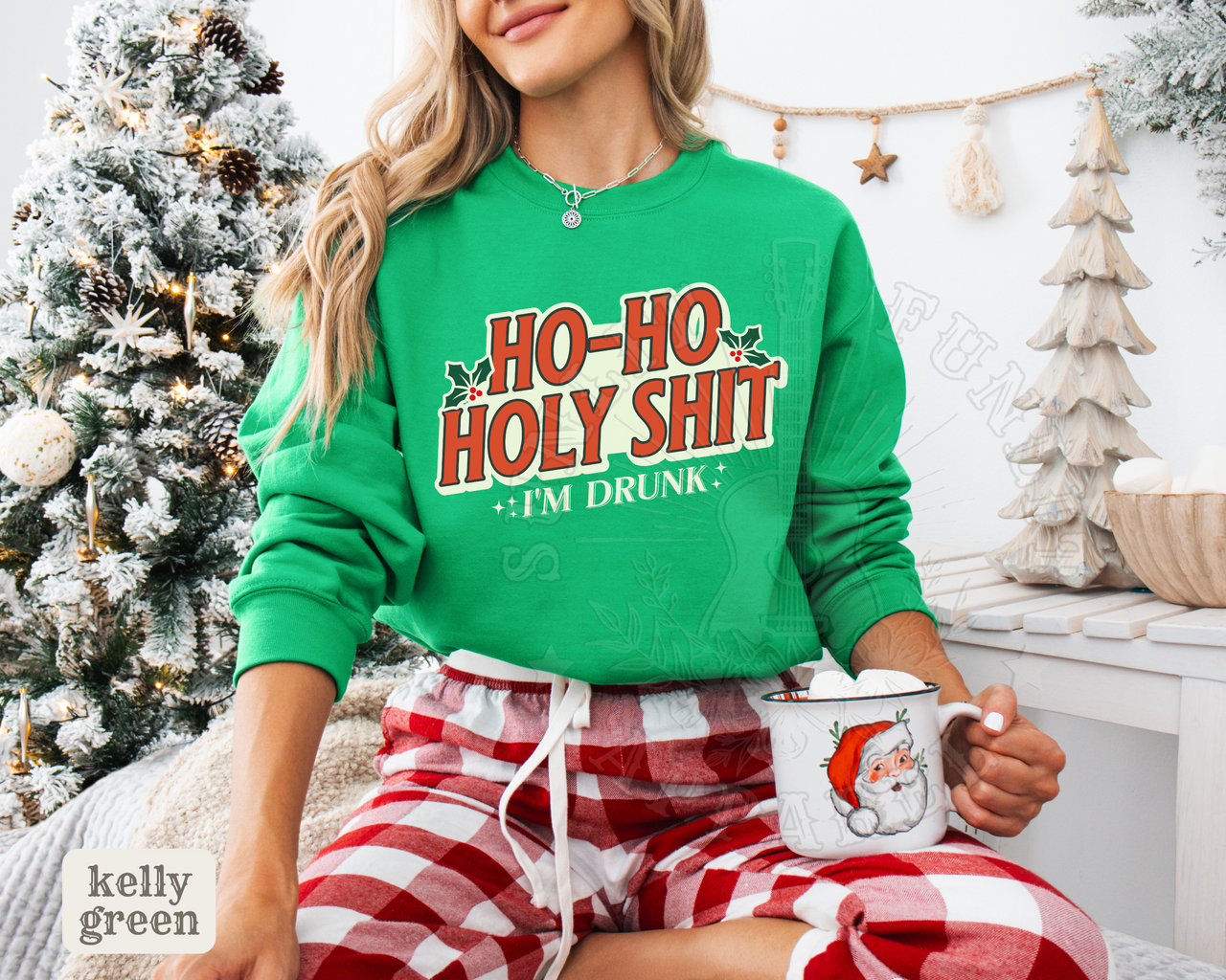 A woman wearing a green sweatshirt with the text "HOO-HO HOLY SHIT I'M DRUNK" is sitting in front of a Christmas tree. She is holding a mug with a Santa Claus design.