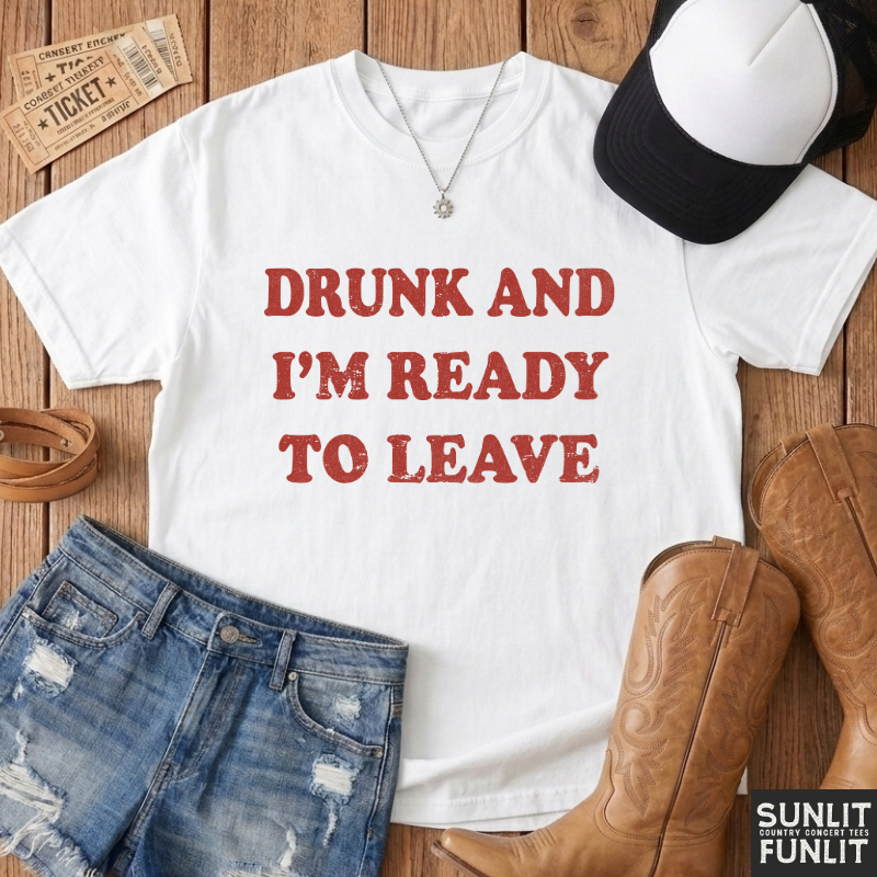 A white graphic t-shirt with the text 'DRUNK AND I'M READY TO LEAVE' in distressed red lettering, styled in a flat lay on a wooden background with jeans, cowboy boots, a baseball cap, and tickets.