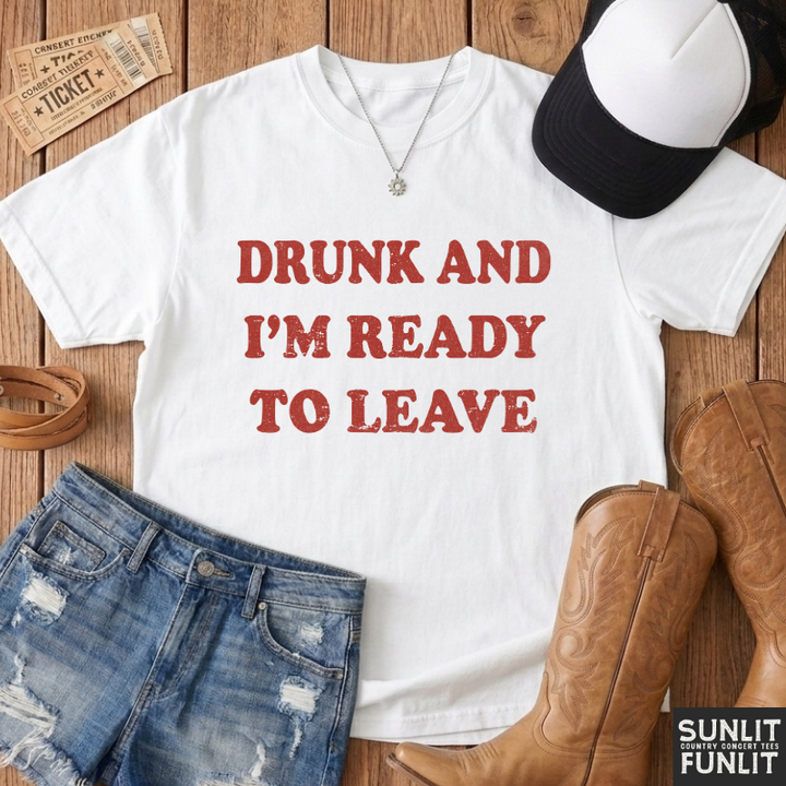 A white graphic t-shirt with the text 'DRUNK AND I'M READY TO LEAVE' in distressed red lettering, styled in a flat lay on a wooden background with jeans, cowboy boots, a baseball cap, and tickets.