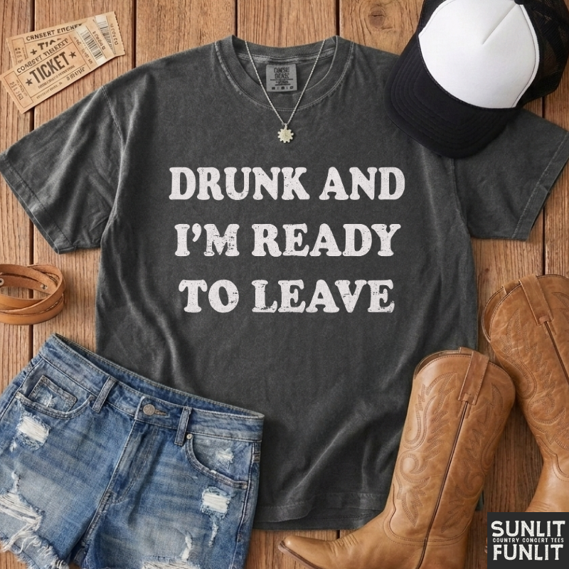 A dark charcoal gray graphic t-shirt with the text 'DRUNK AND I'M READY TO LEAVE' in distressed white lettering, styled in a flat-lay arrangement on a wooden background with jeans, cowboy boots, a cap, and a ticket.