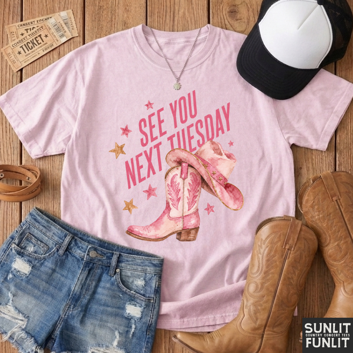 A light pink graphic t-shirt featuring the text 'SEE YOU NEXT TUESDAY' in pink with cowboy boots and stars, styled in a flat lay with denim shorts, a black and white cap, brown cowboy boots, and tickets on a wooden background. The brand logo 'SUNLIT WITH FUNLIT' is visible in the bottom right corner.
