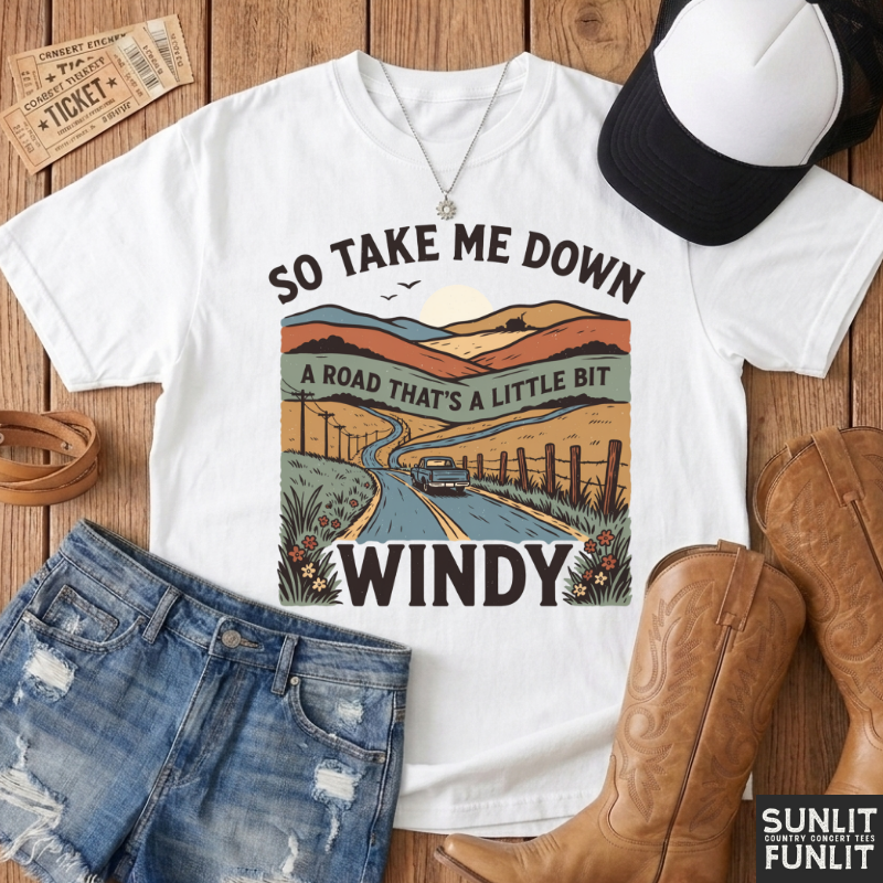 A white graphic T-shirt laid flat on a rustic wood surface, styled with distressed denim shorts, brown cowboy boots, a black-and-white baseball cap, and small ticket props. The shirt graphic shows a winding road through a landscape with the text 'SO TAKE ME DOWN A ROAD THAT'S A LITTLE BIT WINDY.