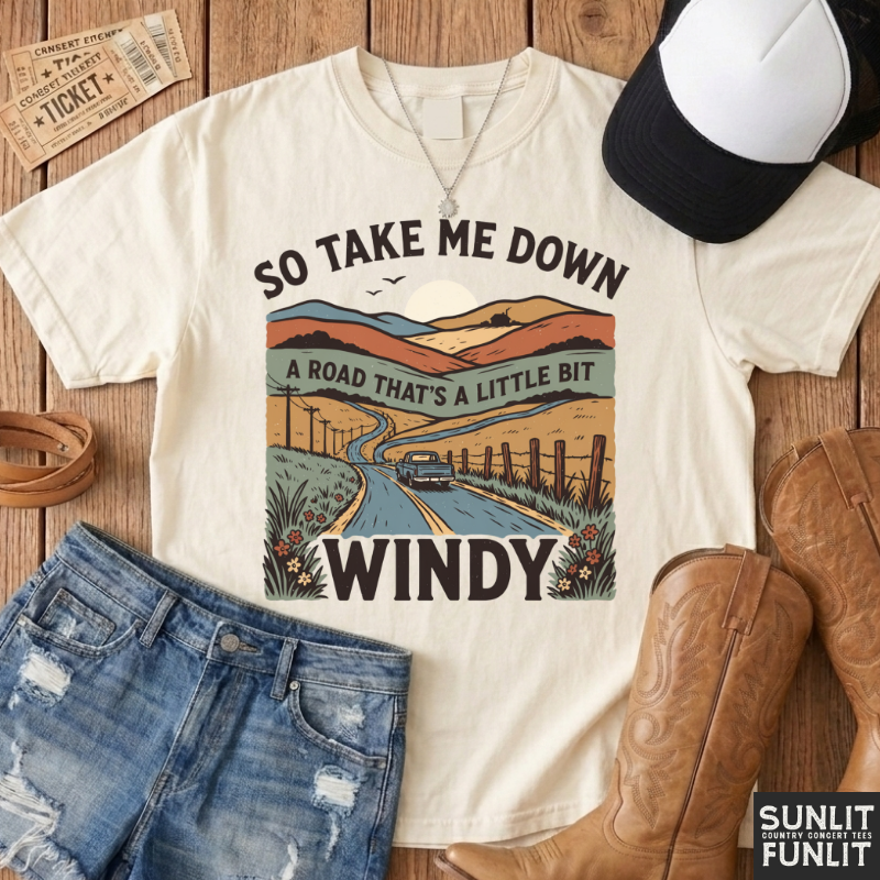 An off-white graphic t-shirt laid flat on a wooden surface, styled with a black and white baseball cap, blue jeans, brown cowboy boots, and vintage-style ticket props. The t-shirt features a retro illustration of a winding road through a mountain landscape with the text 'SO TAKE ME DOWN A ROAD THAT'S A LITTLE BIT WINDY'.