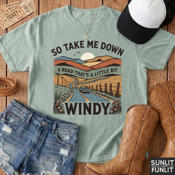 A rust-orange graphic t-shirt styled in a flat lay with denim shorts, cowboy boots, a black and white cap, and vintage-style tickets on a wooden background. The t-shirt features a retro landscape graphic with a winding road, fence, and the text 'SO TAKE ME DOWN A ROAD THAT'S A LITTLE BIT WINDY'.