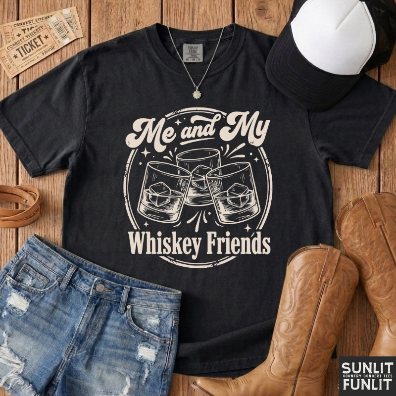 A black graphic t-shirt with a white 'Me and My Whiskey Friends' print, styled in a flat-lay with denim shorts, cowboy boots, a baseball cap, and a vintage ticket on a wooden background.