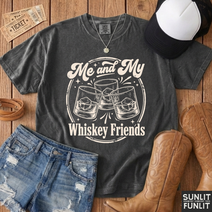 A dark gray graphic t-shirt with a white western-style print reading 'Me and My Whiskey Friends' with clinking glasses, styled in a flat lay on a wooden background with jeans, cowboy boots, a cap, and a ticket.