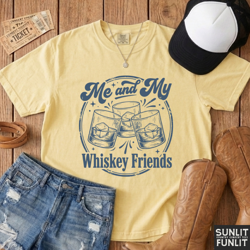 A pale yellow graphic T-shirt laid flat on a rustic wood surface, styled with denim shorts, a black-and-white cap, brown cowboy boots, and a necklace. The shirt reads 'Me and My Whiskey Friends' with an illustration of three clinking whiskey glasses.