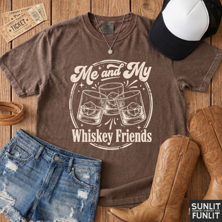 A brown graphic t-shirt with the text 'Me and My Whiskey Friends' and an illustration of three clinking glasses, styled in a flat lay with denim shorts, cowboy boots, a baseball cap, and a leather belt on a wooden background.