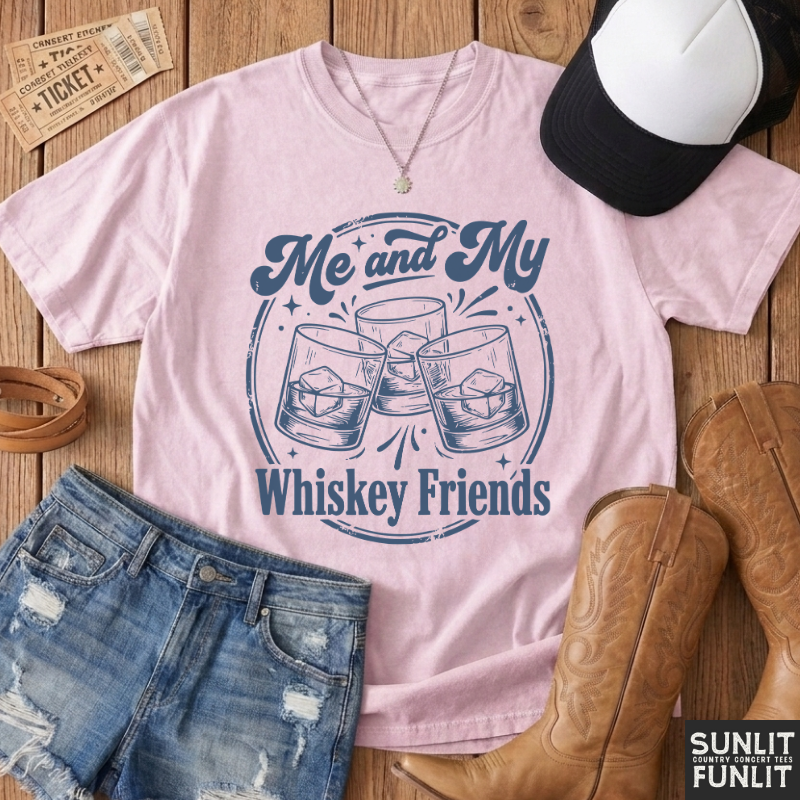 A light pink graphic t-shirt featuring the text 'Me and My Whiskey Friends' in navy blue, styled in a flat-lay arrangement with denim shorts, cowboy boots, a baseball cap, and a necklace on a wooden background.