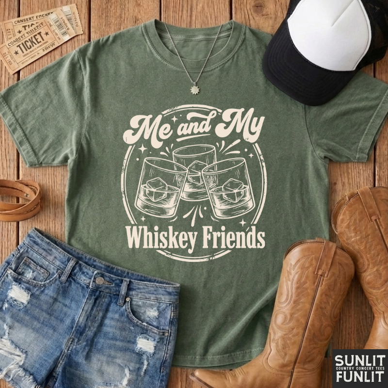 An olive green graphic t-shirt with a white print reading 'Me and My Whiskey Friends' with clinking glasses, styled in a flat-lay on a wooden background with jeans, cowboy boots, a cap, and a ticket.