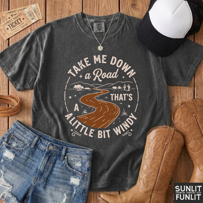 A dark gray graphic t-shirt featuring a western-style design of a winding road through a landscape with the text 'TAKE ME DOWN a Road THAT'S A LITTLE BIT WINDY'. The shirt is styled in a flat-lay arrangement on a wooden background with denim shorts, cowboy boots, a baseball cap, and a ticket.