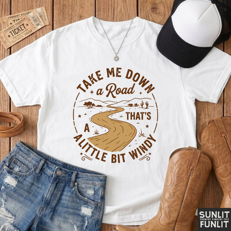 A white graphic t-shirt with a brown circular road design and the text 'TAKE ME DOWN a Road THAT'S A LITTLE BIT WINDY', styled in a flat lay on a wooden surface with denim shorts, cowboy boots, a black and white cap, a necklace, and a ticket stub. A 'SUNLIT CRAFTED FUNLIT' logo is visible in the bottom right corner.