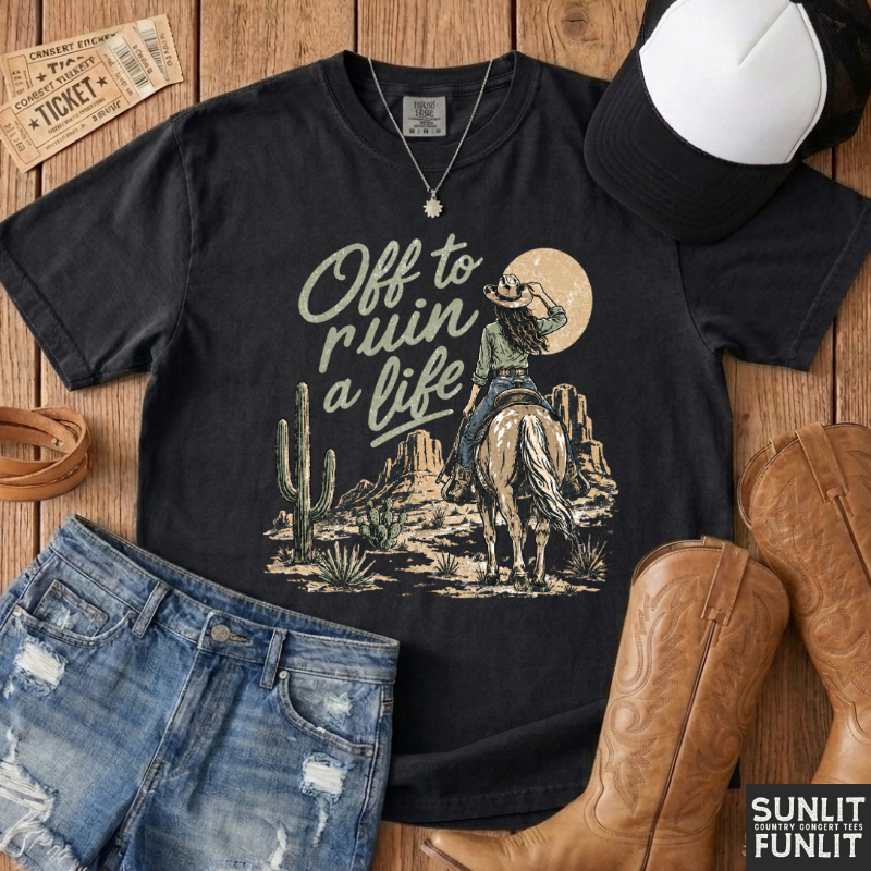 A black graphic t-shirt featuring a western cowboy design with the text 'Off to ruin a life' and a desert landscape, styled in a flat lay with denim shorts, cowboy boots, a baseball cap, and a necklace on a wooden background.