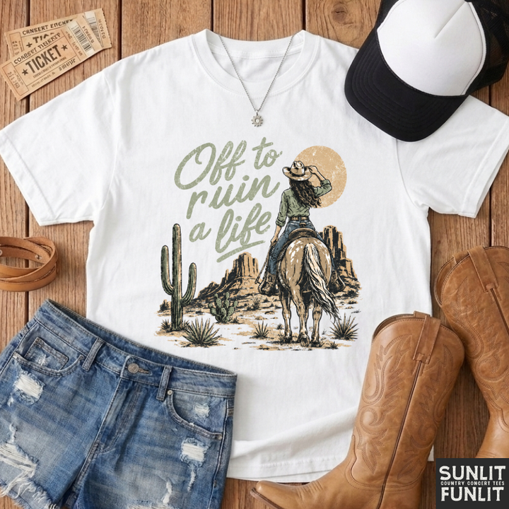 A white graphic T-shirt laid flat on a wooden surface with styled western accessories around it, including a black-and-white cap, denim shorts, brown cowboy boots, and ticket stubs. The shirt graphic reads “Off to ruin a life” with a cowboy on a horse, cactus, and desert rock scene.
