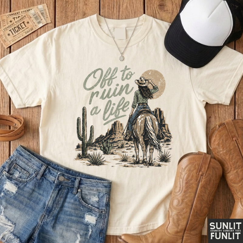An off-white graphic t-shirt featuring a western design of a cowboy on a horse with the text 'Off to ruin a life', styled in a flat lay with denim shorts, cowboy boots, a baseball cap, and a ticket on a wooden background.