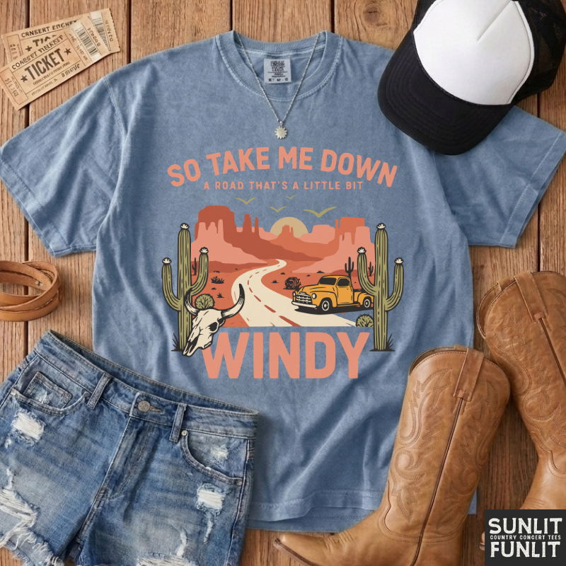 A styled flat-lay of a light blue graphic t-shirt with a desert road trip print, paired with distressed denim shorts, brown cowboy boots, a black and white baseball cap, and a leather belt on a wooden background. The brand logo 'SUNLIT FUNLIT' is visible in the bottom right corner.