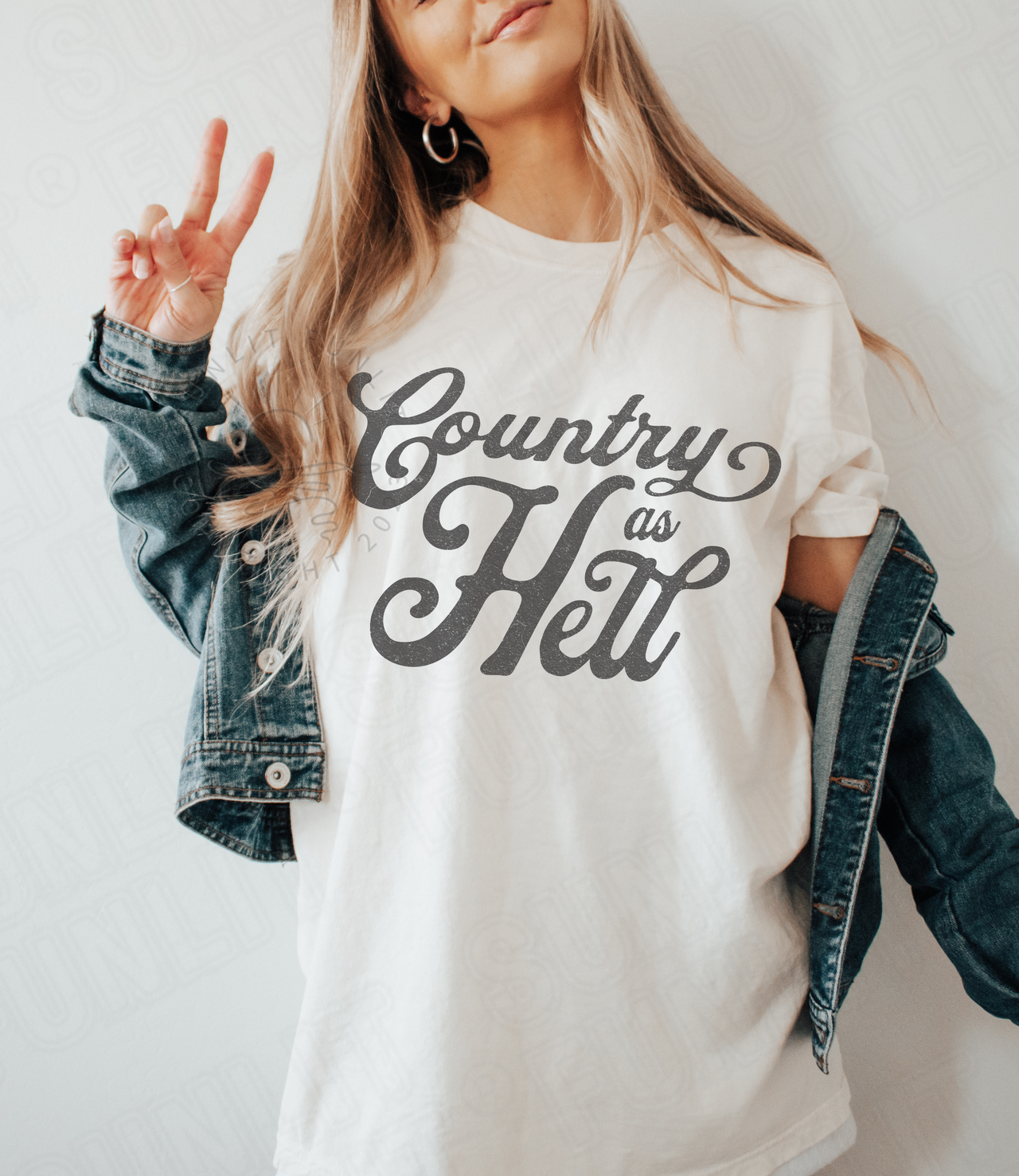 A woman wearing a white t-shirt with the text "Country as Hell" and a denim jacket, making a peace sign gesture.