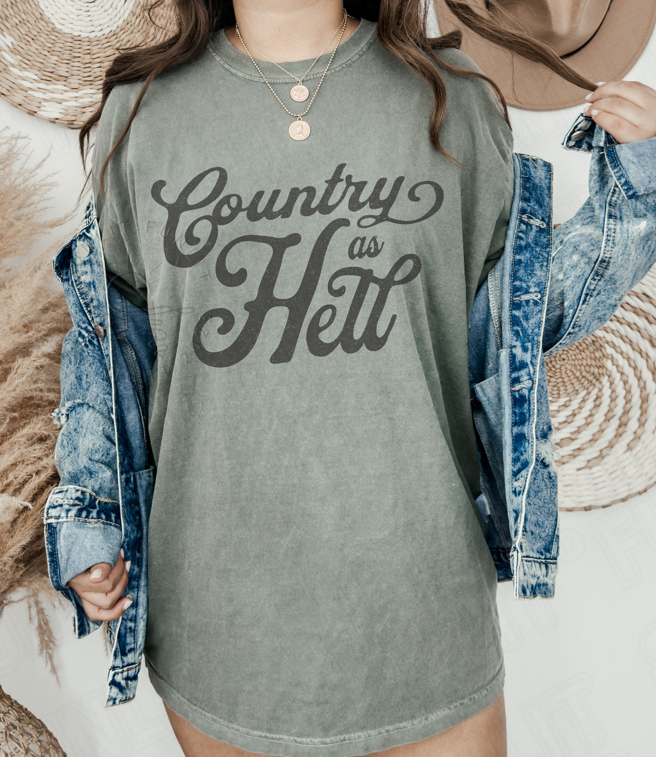 A person wearing a gray t-shirt with the text "Country as Hell" printed on it, paired with a denim jacket and a hat.