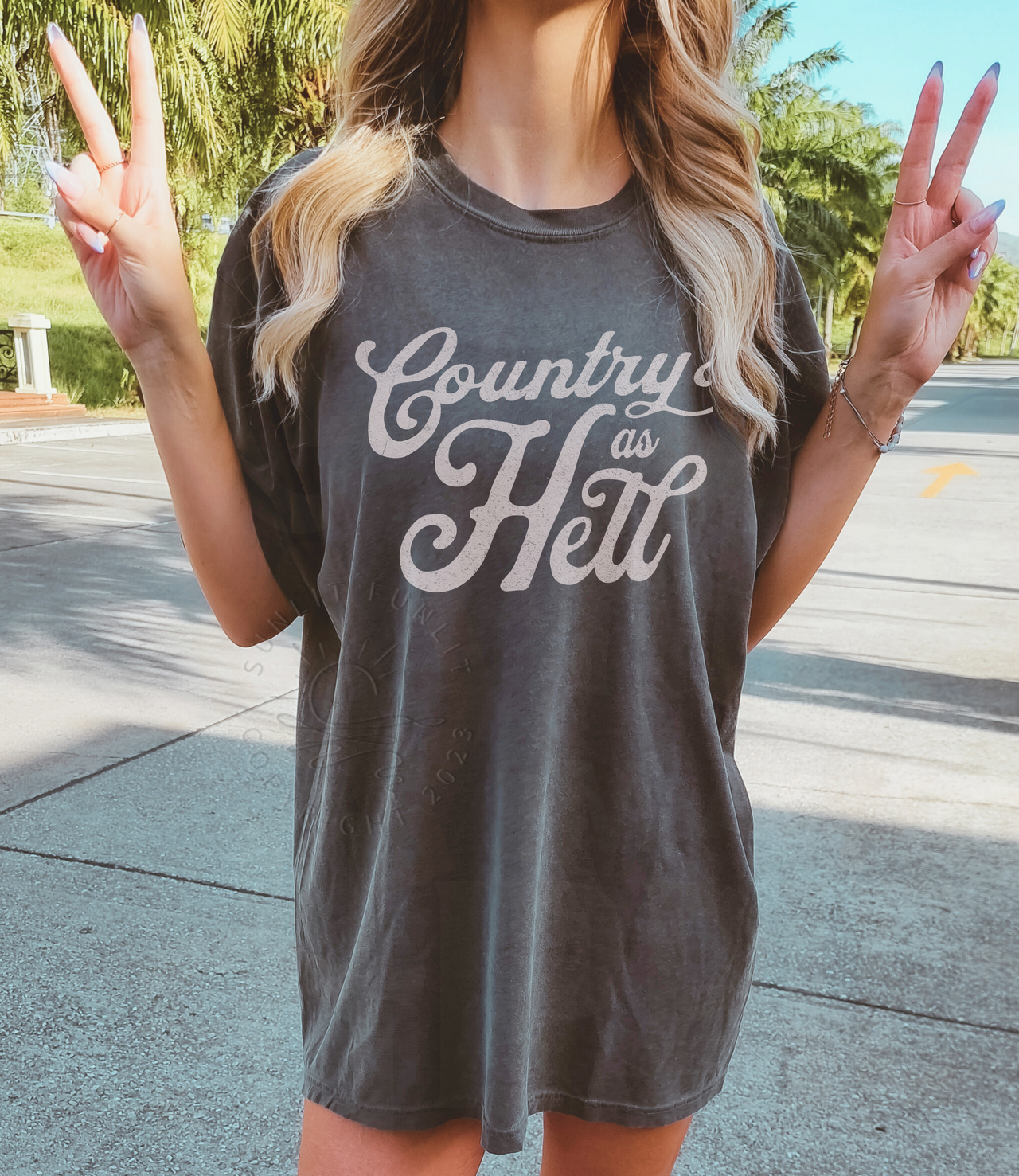 A woman wearing a grey t-shirt with the text "Country as Hell" on it, making a peace sign with her hands.