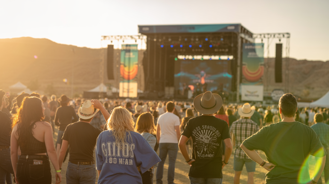 People at a music festival with a stage in the background during sunset.