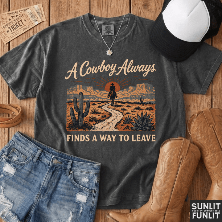 A dark heather gray graphic t-shirt featuring a western desert scene with the text 'A Cowboy Always FINDS A WAY TO LEAVE'. The shirt is styled in a flat lay on a wooden surface with denim shorts, a black and white baseball cap, tan cowboy boots, and a necklace.