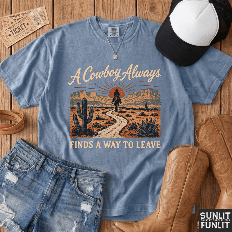 A light blue graphic t-shirt laid flat on a wooden surface, styled with a black and white baseball cap, denim shorts, tan cowboy boots, and vintage-style tickets. The t-shirt features a western desert landscape graphic with the text 'A Cowboy Always FINDS A WAY TO LEAVE'.