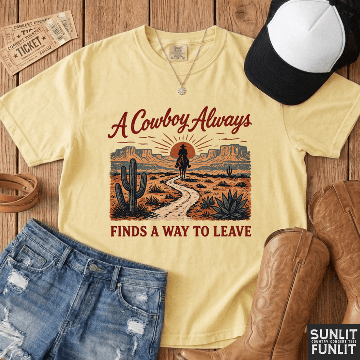 A pale yellow graphic T-shirt laid flat on a rustic wood surface, surrounded by denim shorts, a black-and-white cap, cowboy boots, tickets, and a necklace. The shirt graphic reads “A Cowboy Always Finds A WAY TO LEAVE” with a desert scene and rider illustration.