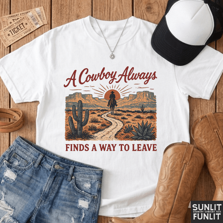 A white graphic t-shirt with a western desert design, styled in a flat lay with blue jeans, a black and white baseball cap, brown cowboy boots, a leather belt, and vintage-style tickets on a wooden background. The t-shirt graphic reads 'A Cowboy Always FINDS A WAY TO LEAVE'.