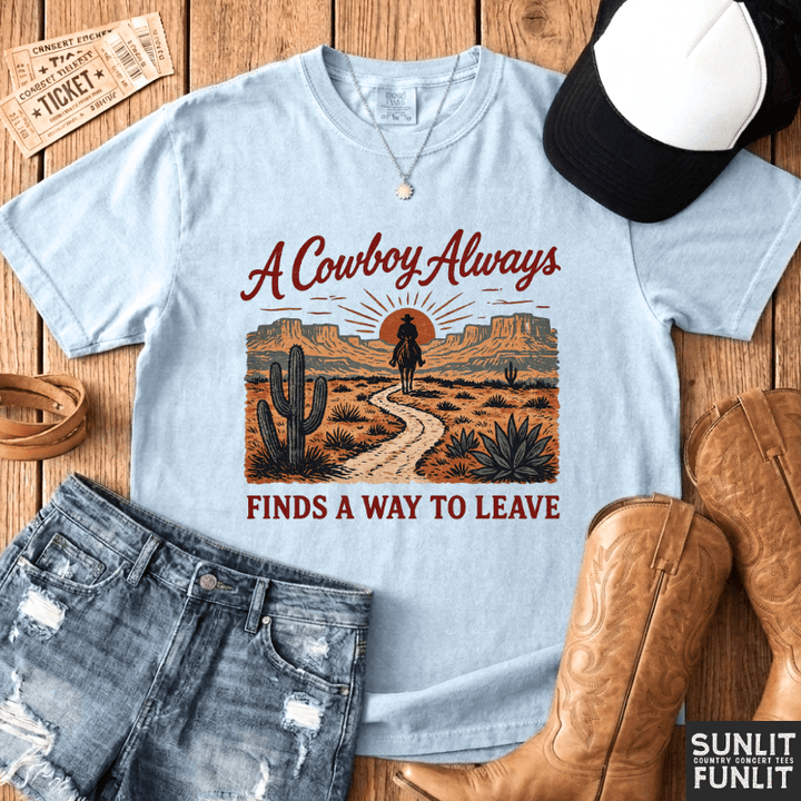 A light blue graphic t-shirt laid flat on a wooden surface, styled with a black and white baseball cap, denim shorts, brown cowboy boots, and a cowboy ticket. The t-shirt features a desert landscape graphic with a cowboy silhouette and the text 'A Cowboy Always FINDS A WAY TO LEAVE'.