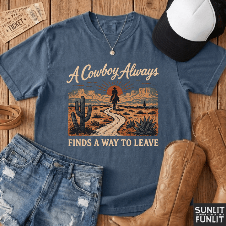 A blue graphic t-shirt laid flat on a wooden surface with styled western accessories, including a black-and-white cap, denim shorts, brown cowboy boots, and a ticket. The shirt reads 'A Cowboy Always FINDS A WAY TO LEAVE' with a desert landscape illustration showing a cowboy riding along a path.