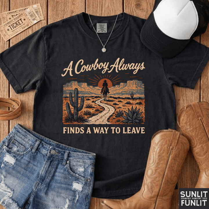 A black graphic t-shirt featuring a western desert scene with the text 'A Cowboy Always FINDS A WAY TO LEAVE', styled in a flat lay with denim shorts, cowboy boots, a baseball cap, and a necklace on a wooden background.