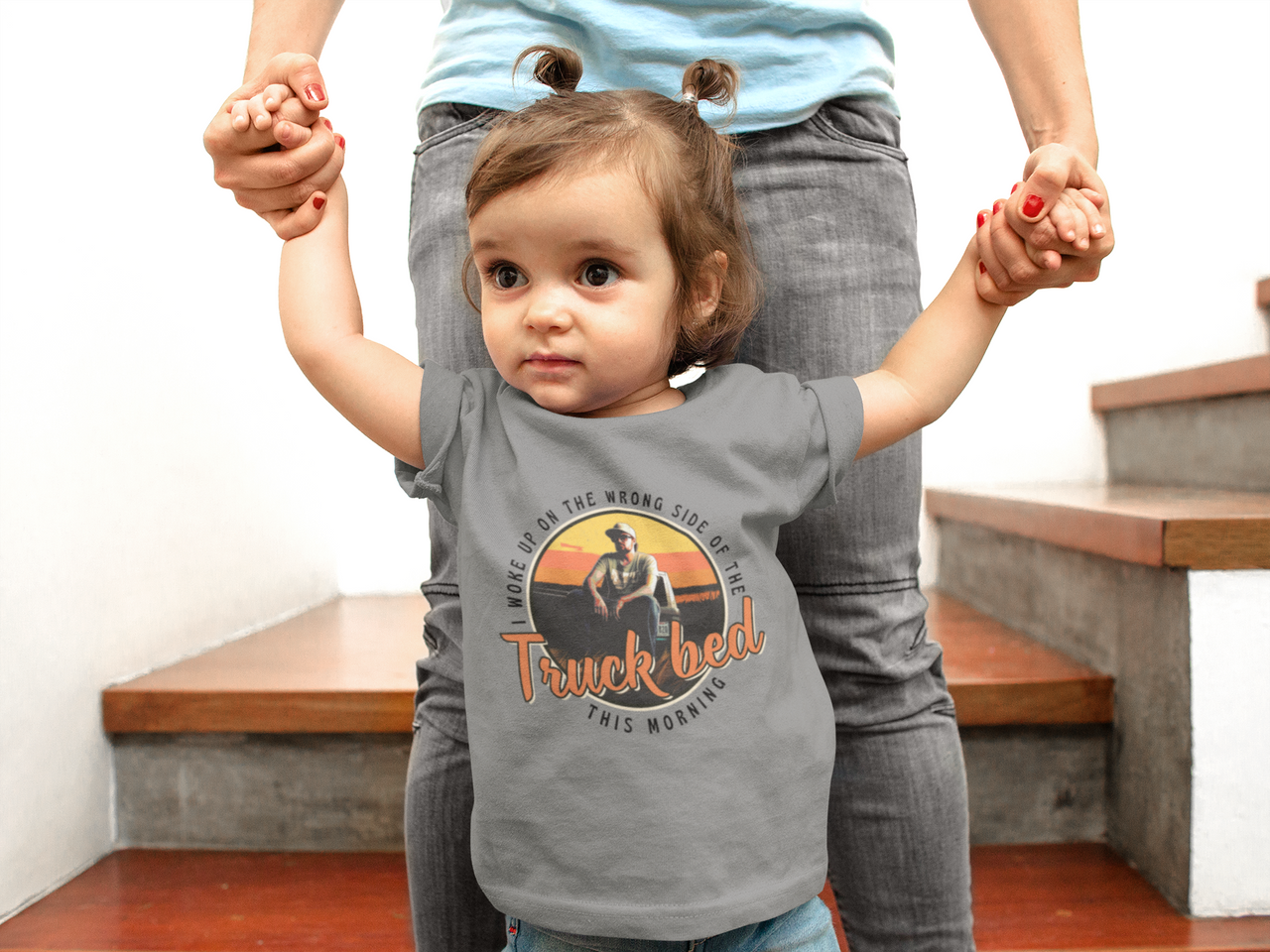 A young child wearing a gray t-shirt with a graphic design and text, standing in front of an adult who is holding their hands.