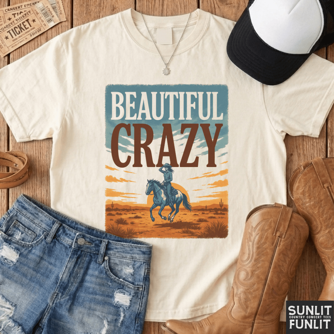 A cream-colored graphic t-shirt featuring a western-themed print that reads 'BEAUTIFUL CRAZY' with an illustration of a cowboy on a horse. The shirt is styled in a flat-lay arrangement with blue jeans, brown cowboy boots, a black and white baseball cap, a necklace, and a vintage ticket on a wooden surface.