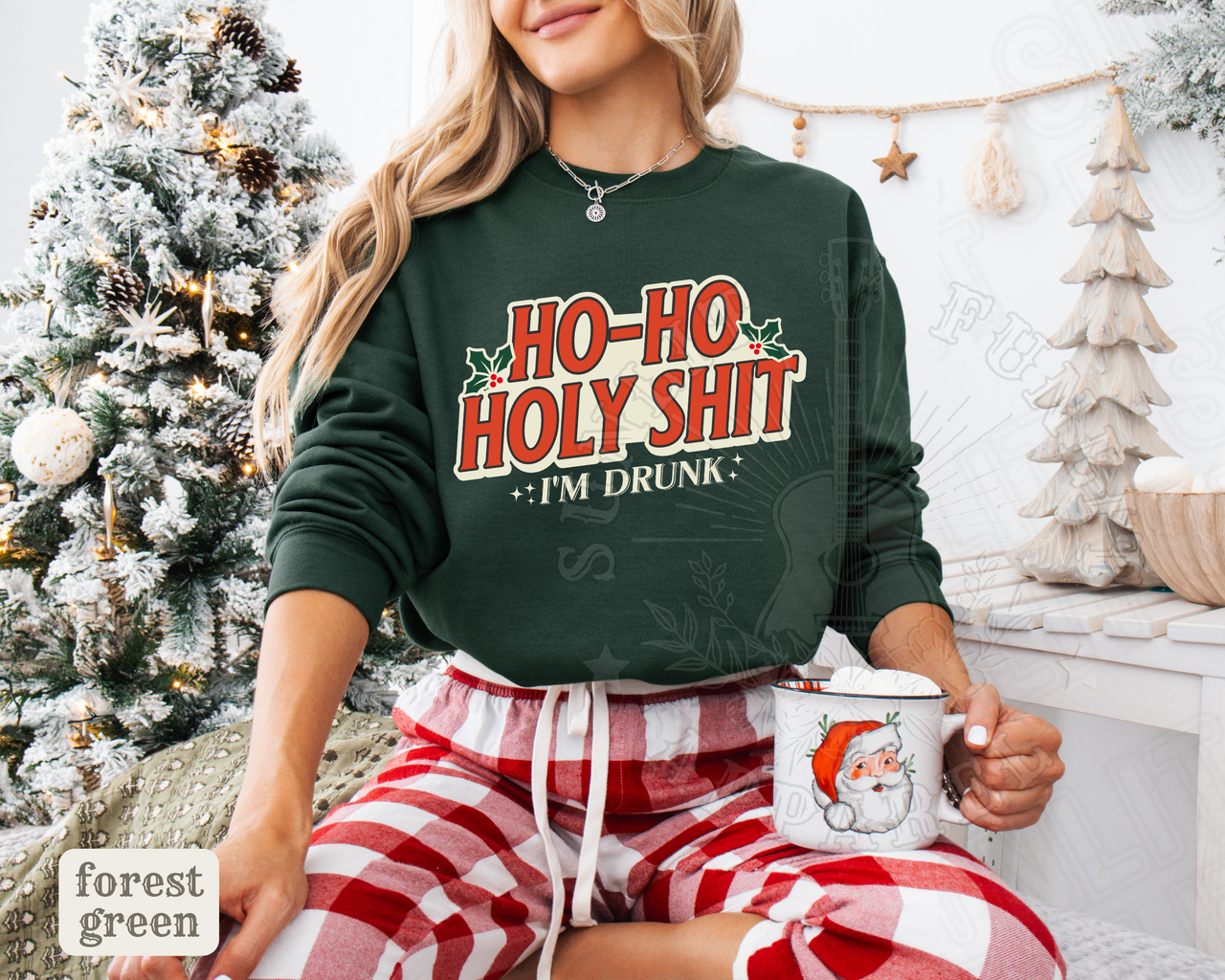 A woman wearing a green sweatshirt with the text "HO-HO HOLY SHIT I'M DRUNK" is sitting in front of a Christmas tree. She is holding a mug with a Santa Claus image on it.