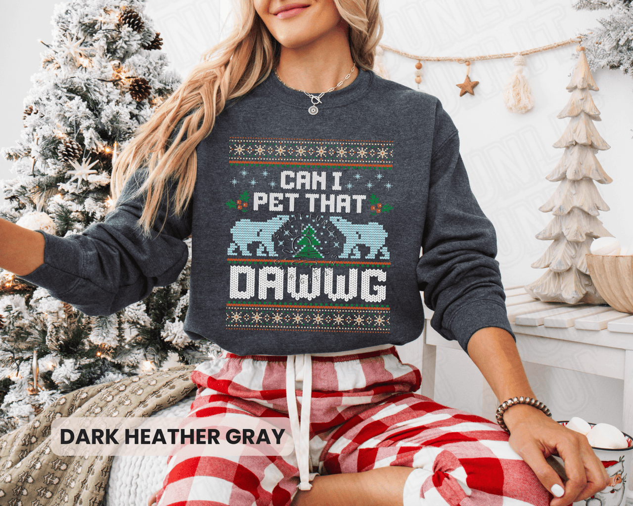 A woman wearing a gray sweatshirt with a Christmas-themed design and text, sitting in front of a Christmas tree.