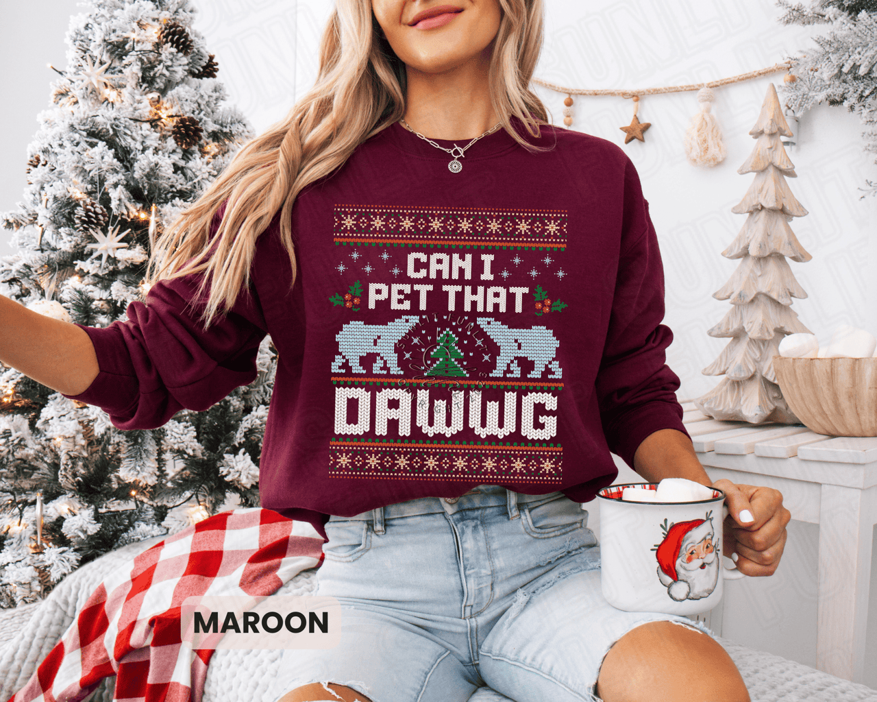 A woman wearing a burgundy sweater with a Christmas-themed design and text, sitting in front of a Christmas tree and holding a mug.