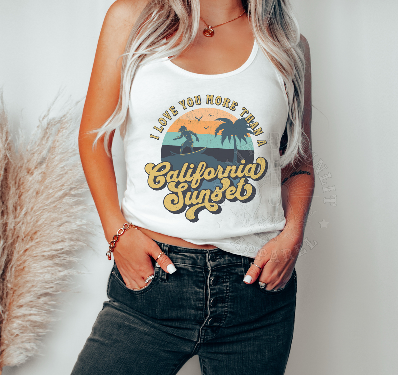 A woman wearing a white tank top with a graphic design that says "I LOVE YOU MORE THAN A CALIFORNIA SUNSETS" and jeans.