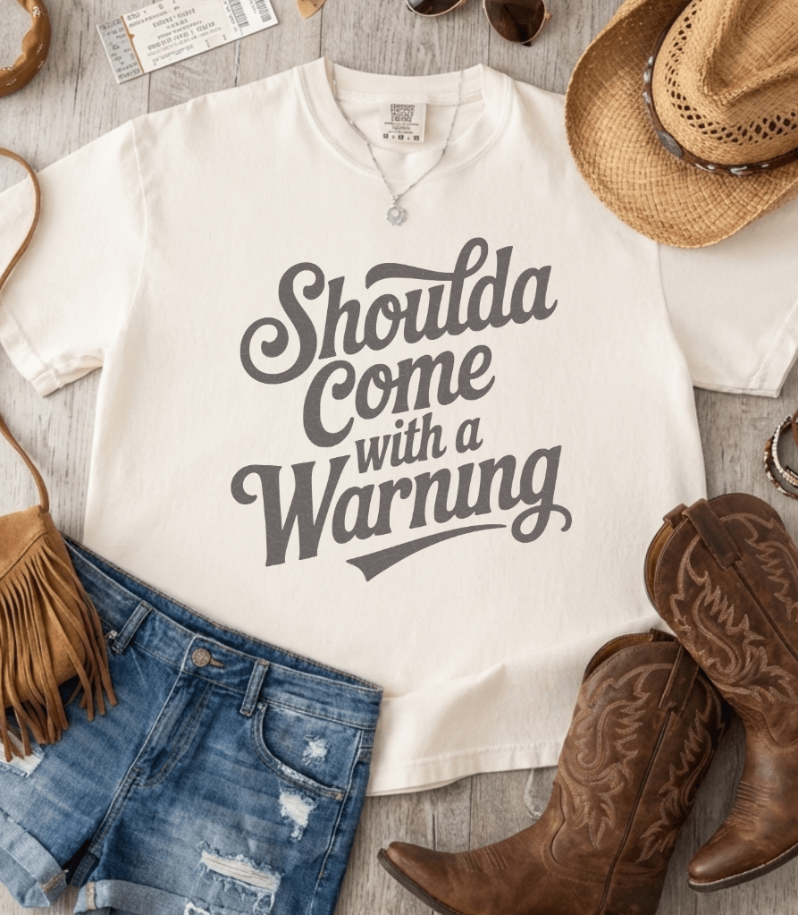 White t-shirt with 'Shoulda Come with a Warning' text, paired with denim shorts, cowboy boots, and a hat on a wooden surface.