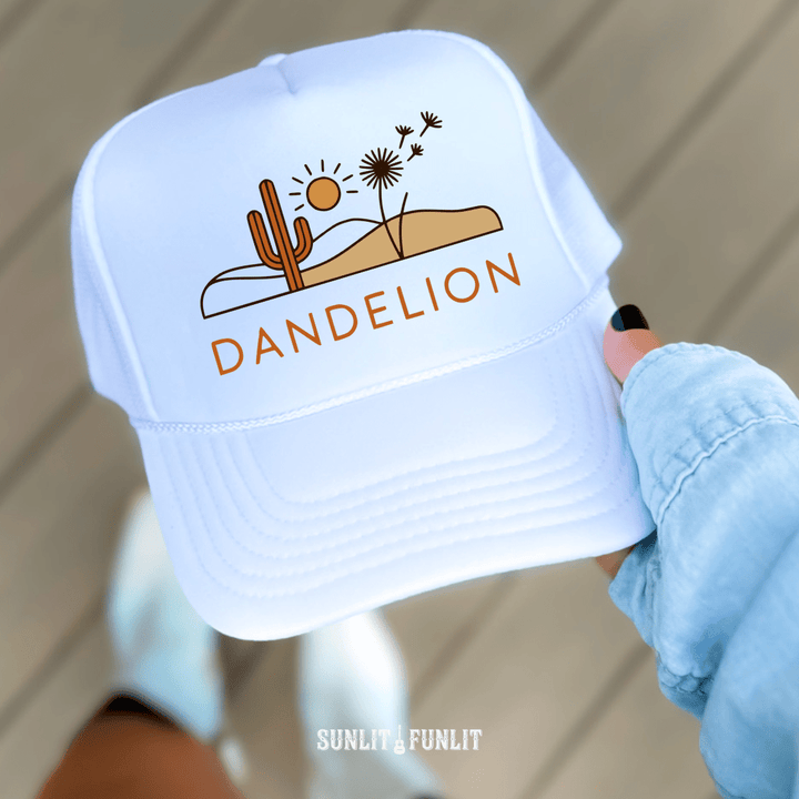 White cap with desert-themed design and 'Dandelion' text held by a person wearing a light blue jacket.