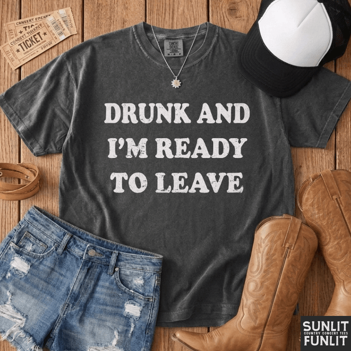 A dark charcoal gray graphic t-shirt with the text 'DRUNK AND I'M READY TO LEAVE' in distressed white lettering, styled in a flat-lay arrangement on a wooden background with jeans, cowboy boots, a cap, and a ticket.