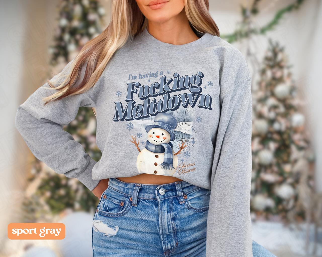 A woman wearing a gray sweatshirt with a snowman graphic and text on it, standing in front of a Christmas tree.