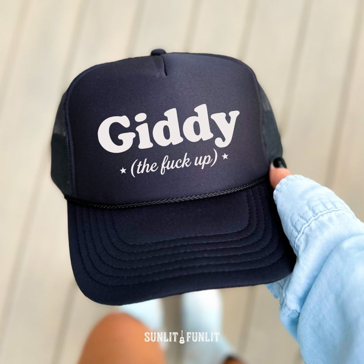 A black trucker hat with white text reading 'Giddy (the fuck up)' held by a hand wearing a light blue denim sleeve, against a light beige paneled background.