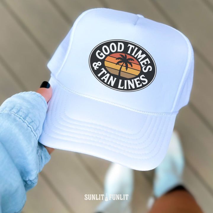 White cap with 'Good Times & Tan Lines' design held by a person wearing a blue jacket.