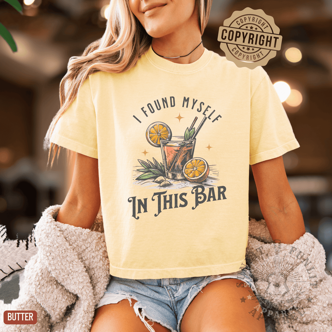 A woman wearing a yellow t-shirt with a graphic design of a cocktail and the text "I FOUND MYself IN THIS BAR" printed on it.