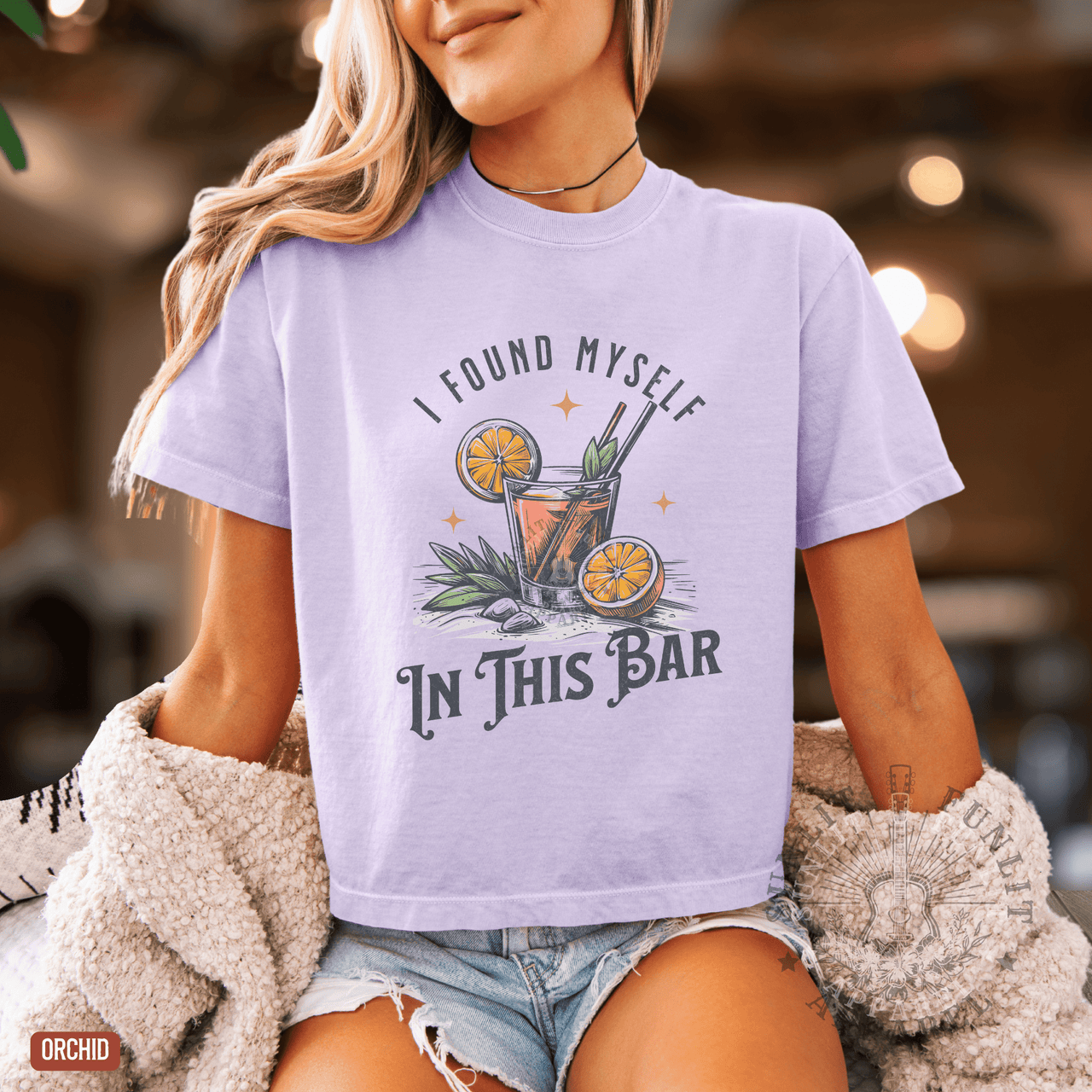 A woman wearing a purple t-shirt with a graphic design of a cocktail and the text "I FOUND MYself IN THIS BAR" printed on it.