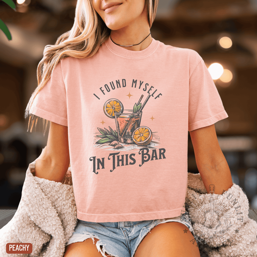A woman wearing a pink t-shirt with a graphic design of a cocktail and the text "I FOUND MYself IN THIS BAR" is shown in the image.