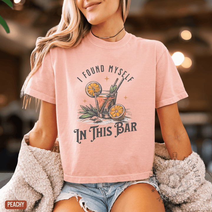 A woman wearing a pink t-shirt with a graphic design of a cocktail and the text "I FOUND MYself IN THIS BAR" is shown in the image.