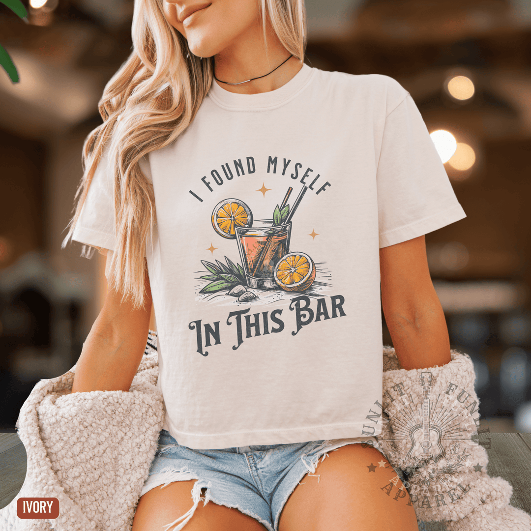 A woman wearing a white t-shirt with a graphic design of a cocktail and the text "I FOUND MYself IN THIS BAR" printed on it.