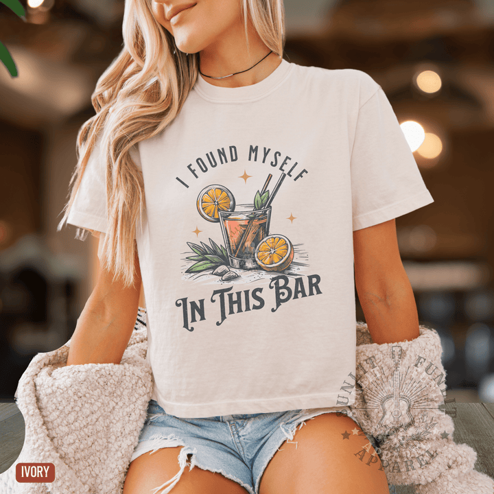 A woman wearing a white t-shirt with a graphic design of a cocktail and the text "I FOUND MYself IN THIS BAR" printed on it.